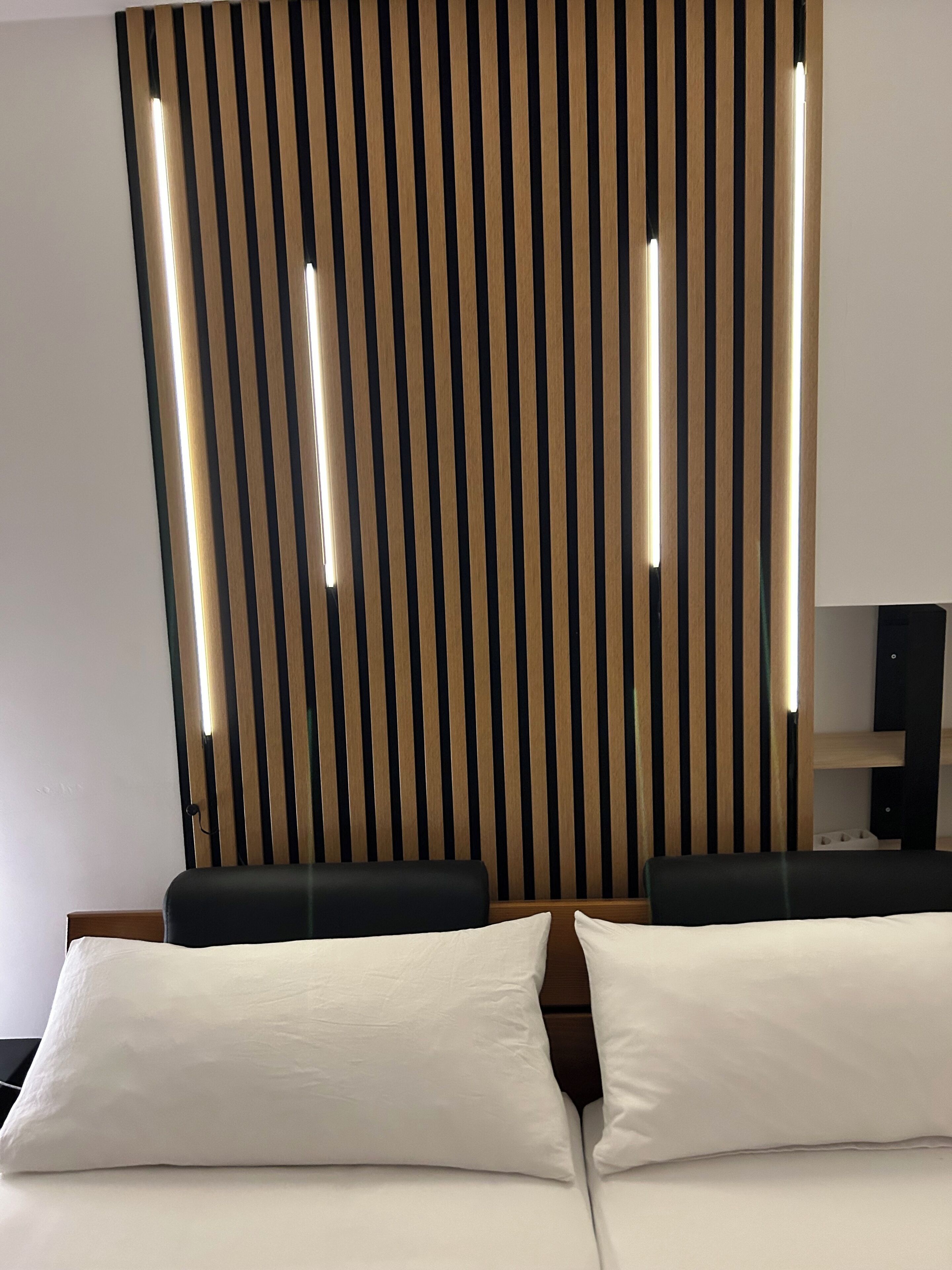 A hotel room with a bed, two pillows, a wooden slat wall, and a speaker.