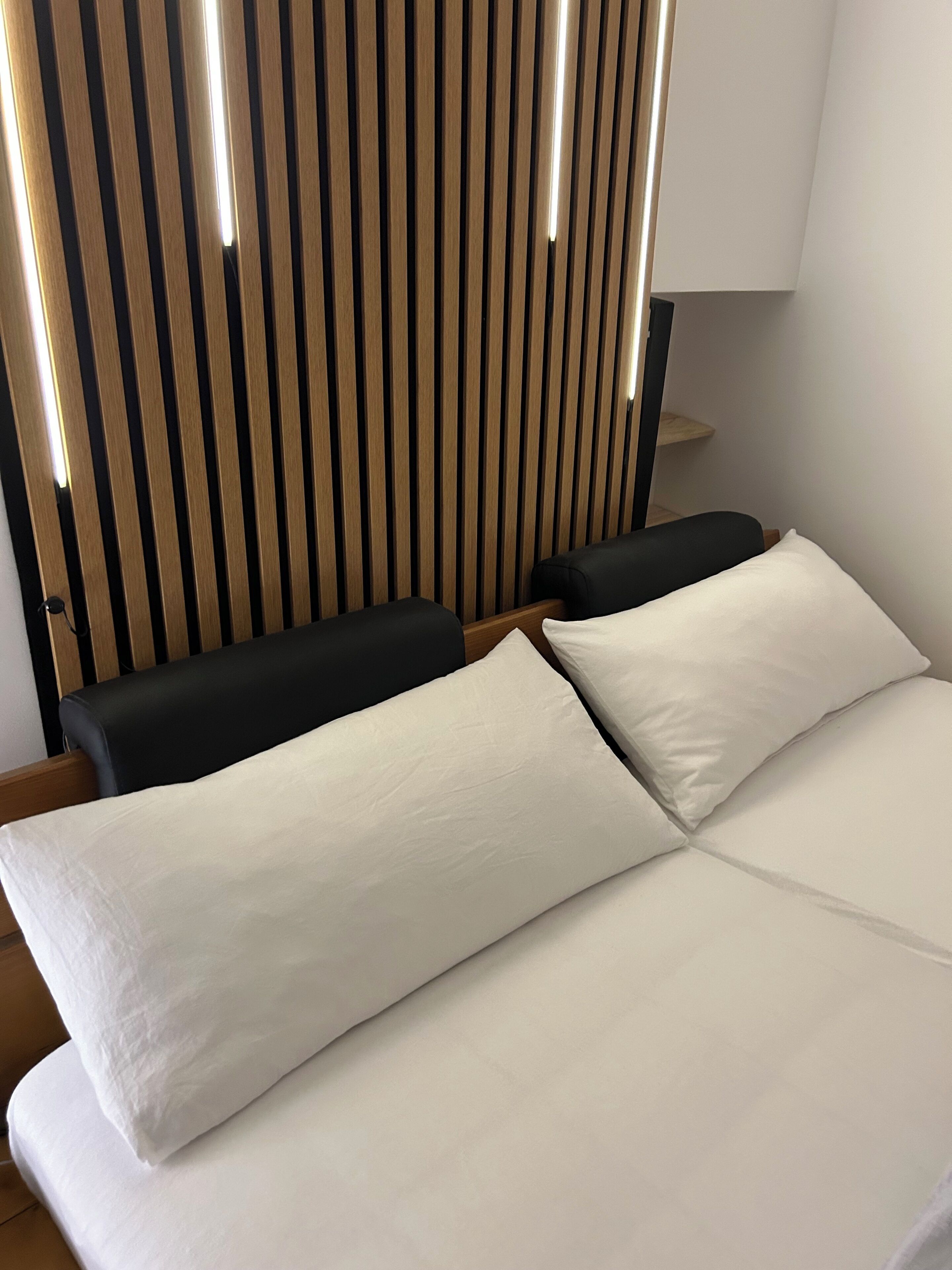 Una cama con ropa blanca, dos sillas negras y un cabecero de madera con listones.