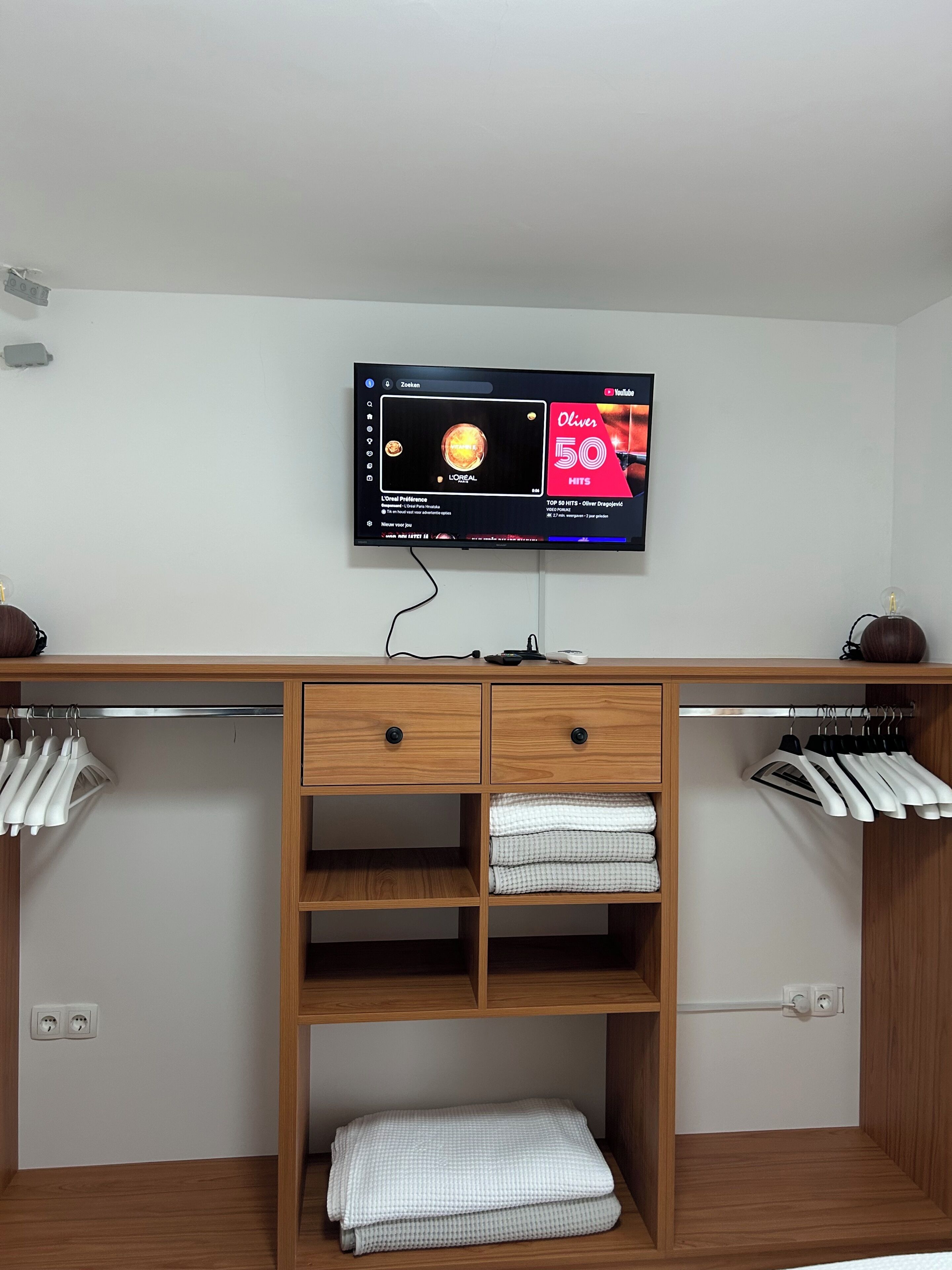 A modern room with a wooden desk, a flat-screen TV mounted above it, and a wardrobe with hanging clothes.