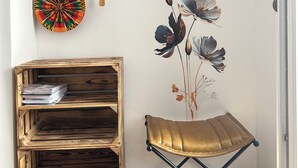 Una habitación de esquina con una estantería de madera, un reloj de pared, un plato decorativo, un mural floral y un taburete plegable.