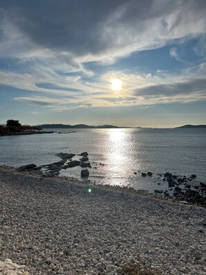 En stenstrand med et roligt hav, fjerne bakker og en klar himmel.