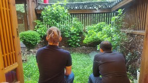 Dos personas sentadas sobre esteras de tatami, frente a un jardín con cerca de madera y follaje.