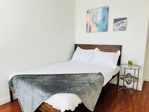 Ein Bett mit grauer Decke, weißen Kissen und einem hölzernen Kopfteil. Es gibt einen kleinen runden Tisch mit Glasplatte und Metallfuß.