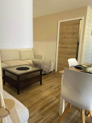 A living room with a sofa, a coffee table, a dining table with chairs, and a wooden door. - Le Bruit des Vagues, an apartment 50 meters from the ocean (Quend)