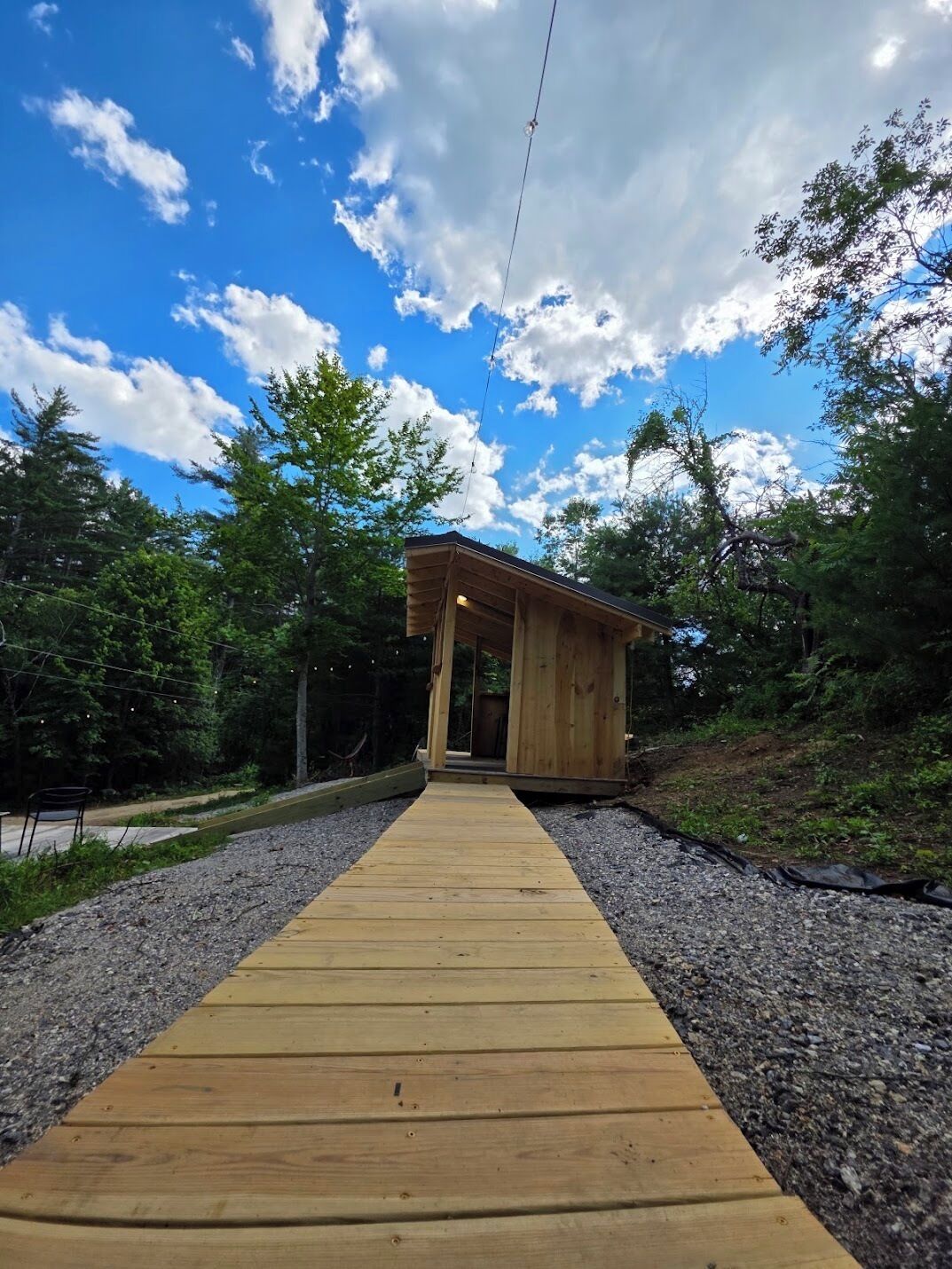 A wooden structure with a covered entrance, surrounded by gravel and trees, under a partly cloudy sky.