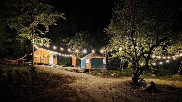 Un terrain de camping avec deux cabanes en bois, un sentier de terre et des guirlandes lumineuses.