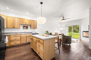 A modern kitchen with wooden cabinets, a central island, and a dining area with a view of the outdoors. - Lower Columbia Landing | 2BR Near Glacier (Columbia Falls)