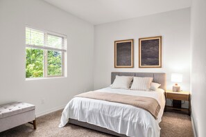 A bedroom with a bed, a nightstand, a lamp, a window with blinds, and two framed pictures on the wall.