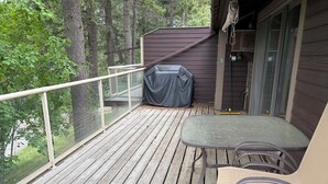 En træterrasse med et overdækket bord og stole, gelænder i glas og et overdækket område med en kost.