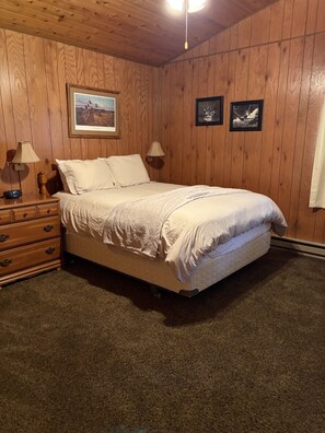 Ein Schlafzimmer mit einer holzverkleideten Wand, einem Bett mit weißer Bettwäsche, zwei gerahmten Bildern, einem Nachttisch mit Lampe und einer Kommode mit Schubladen.