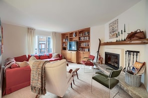 Ein Wohnzimmer mit einem roten Sofa, einem beigen Sessel, einem grünen Stuhl, einem Kamin, einem Bücherregal und einem Fernseher.