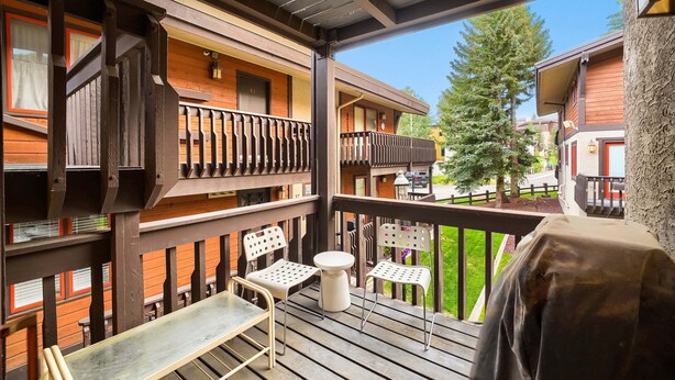 Un balcone in legno con tavolo e sedie, un barbecue e vista su un paesaggio verde e altri edifici.