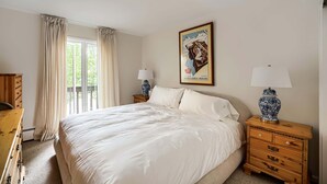 Una camera da letto con un letto grande, una cassettiera in legno, un comodino con una lampada, una finestra con tende e un quadro appeso al muro.