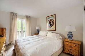 Ein Schlafzimmer mit einem großen Bett, einer Holzkommode, einem Nachttisch mit Lampe, einem Fenster mit Vorhängen und einem gerahmten Bild an der Wand.
