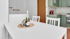 Een witte eettafel met een fles wijn en glazen, een houten stoel, en een keuken met groene kasten en een rode apparatuur.