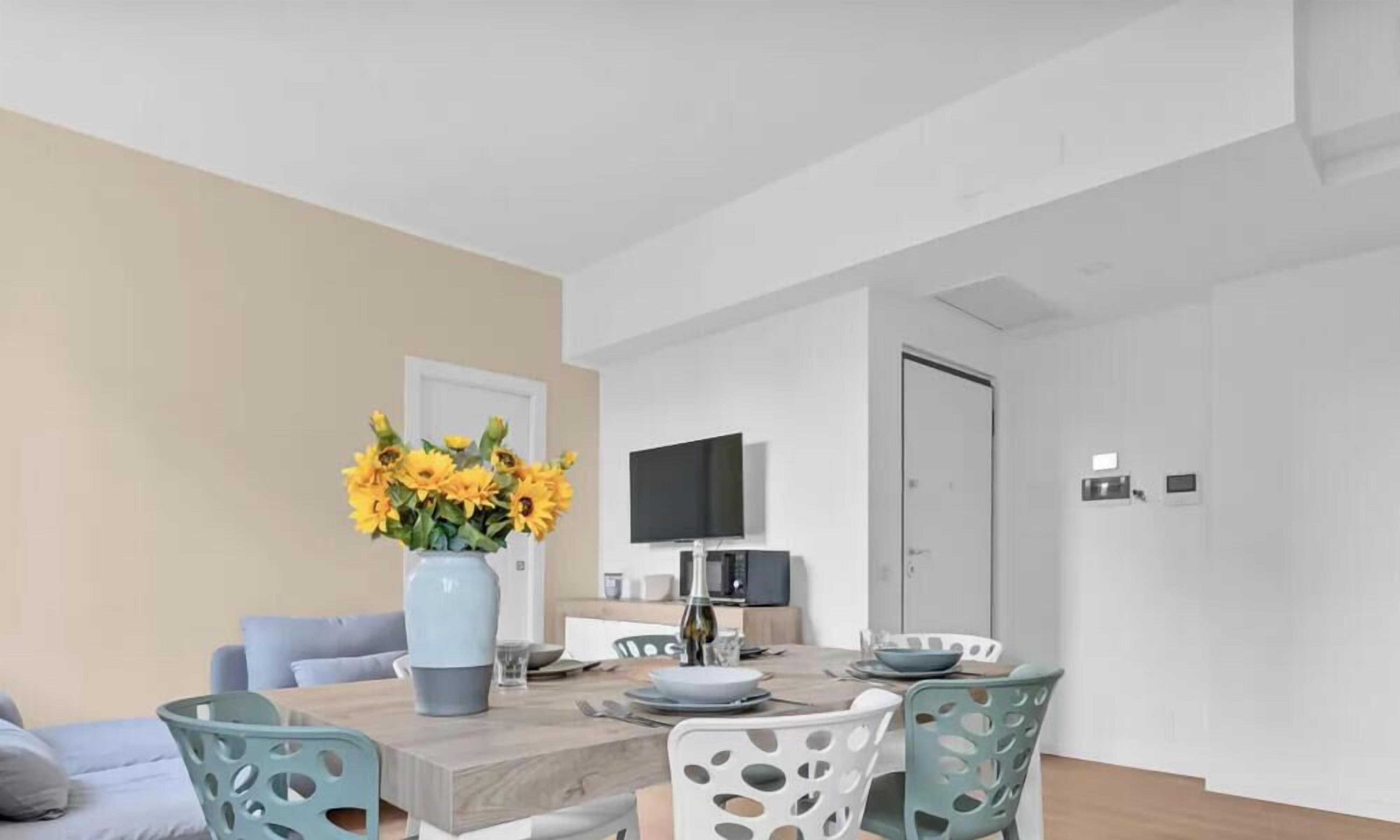 Un espace repas moderne avec une table en bois, des chaises, un vase rempli de tournesols et un téléviseur à écran plat fixé au mur.
