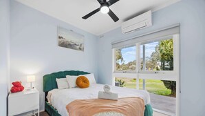Ein Schlafzimmer mit Bett, Nachttisch, Deckenventilator, Klimaanlage, Fenster mit Blick ins Freie und einem Bild an der Wand.
