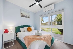 A bedroom with a bed, a nightstand, a ceiling fan, an air conditioning unit, a window with a view of the outdoors, and a painting on the wall.