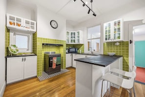 A kitchen with white cabinets, a microwave, a stove, and a clock on the wall. - Brighton Belle — Mid-Century 2BR Home, Split Bath, Walk to Station & Main St (Stawell)