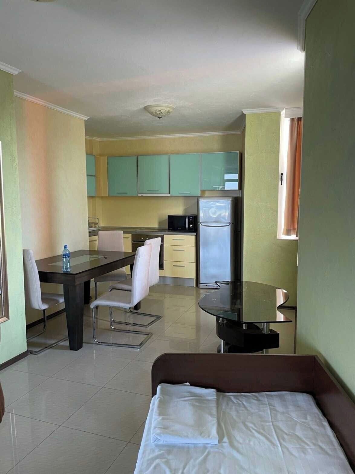 Una habitación de hotel funcional con cama, mesa de comedor con sillas, kitchenette con refrigerador y microondas, y un ventanal con cortinas.