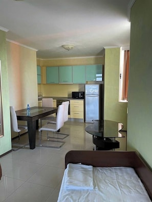 Una habitación de hotel funcional con cama, mesa de comedor con sillas, kitchenette con refrigerador y microondas, y un ventanal con cortinas.
