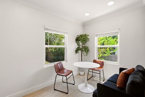 Ein Zimmer mit zwei Fenstern, einem runden Tisch und einem schwarzen Sofa mit braunen Kissen.