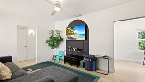 Een woonkamer met een flatscreen televisie aan de muur, een plafondventilator, een bank, een salontafel, een potplant en een bijzettafeltje met een lamp.