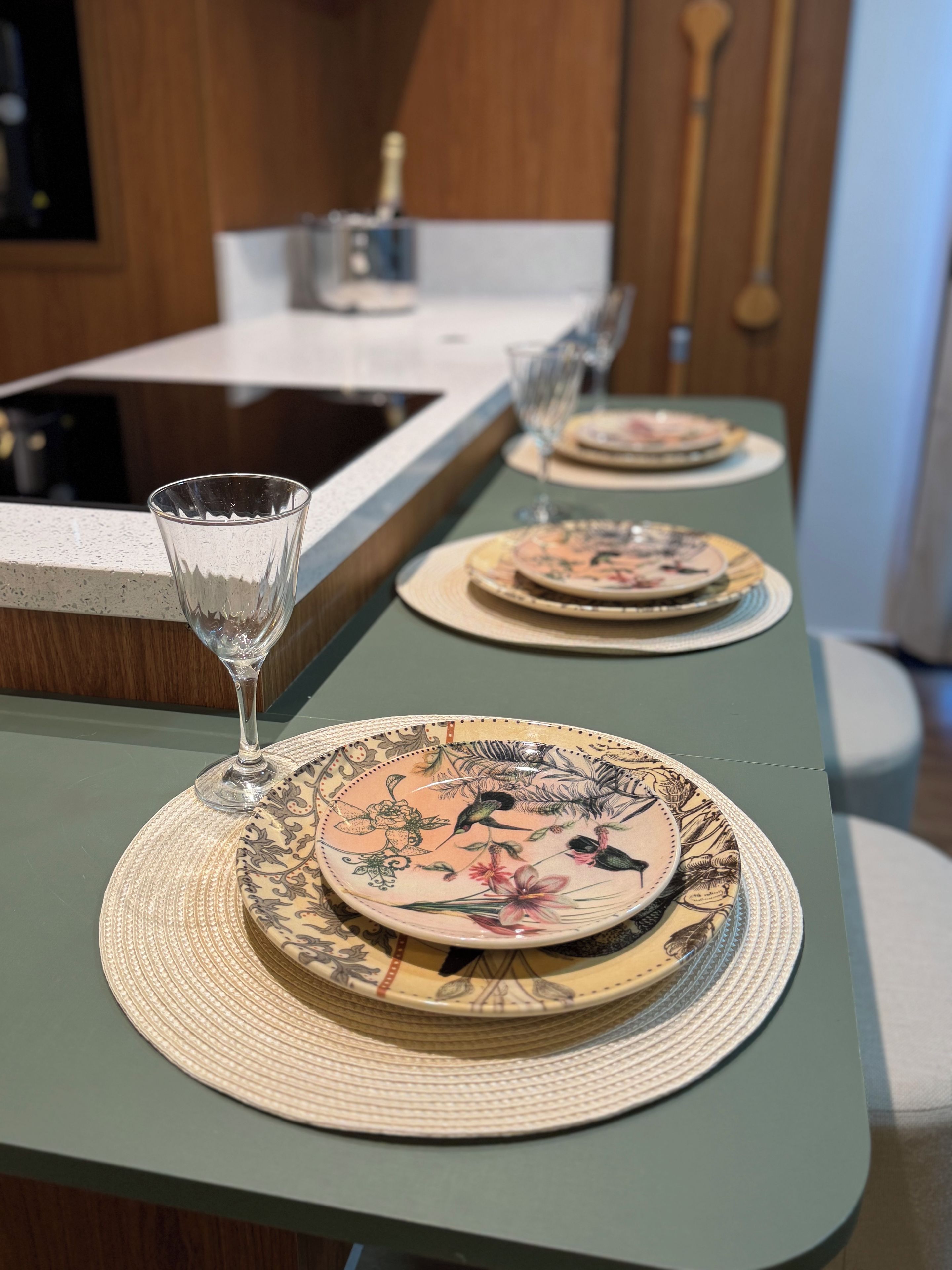 Mesa de jantar com toalha verde, duas travessas com desenhos florais e um copo de vidro transparente.
