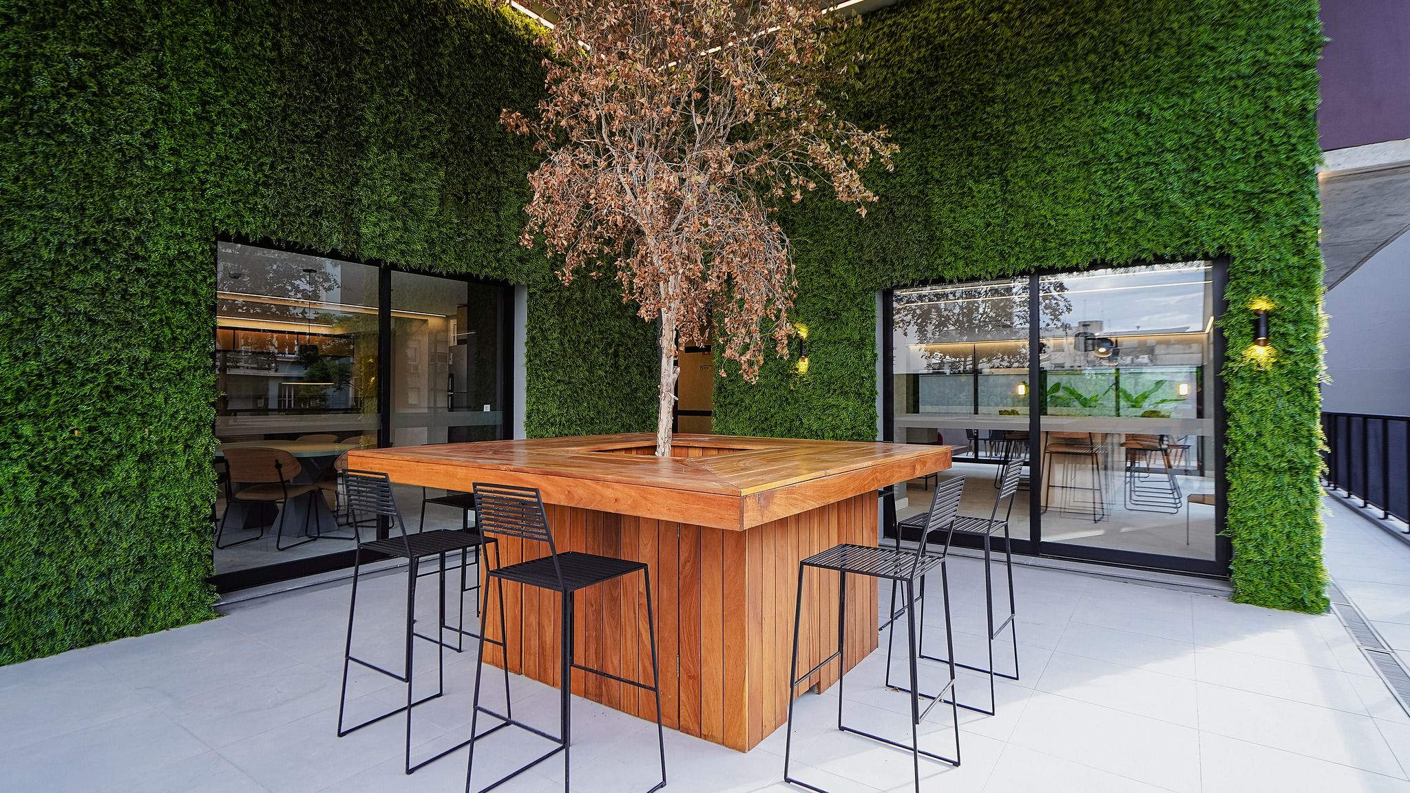 Área de bar ao ar livre com balcão de madeira e banquetas de metal, cercada por paredes verdes exuberantes e amplas janelas.