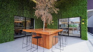 An outdoor bar area with a wooden counter and metal stools, surrounded by lush green walls and large windows.