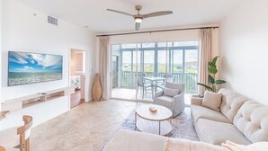 A spacious living room with a large TV, a ceiling fan, a balcony with a table and chairs, and a sofa. - FABULOUS 2B/2B ISLAND CONDO! POOL! WALK TO BEACH! WALK TO EVERYTHING! (Fort Myers Beach)