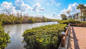 A river with dense vegetation on both sides, a wooden walkway, and buildings in the background. - FABULOUS 2B/2B ISLAND CONDO! POOL! WALK TO BEACH! WALK TO EVERYTHING! (Fort Myers Beach)