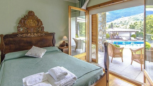 Ein Schlafzimmer mit einem großen Bett, einem Holzkopfstück, Blick auf einen Pool und einem Balkon mit Korbstühlen.