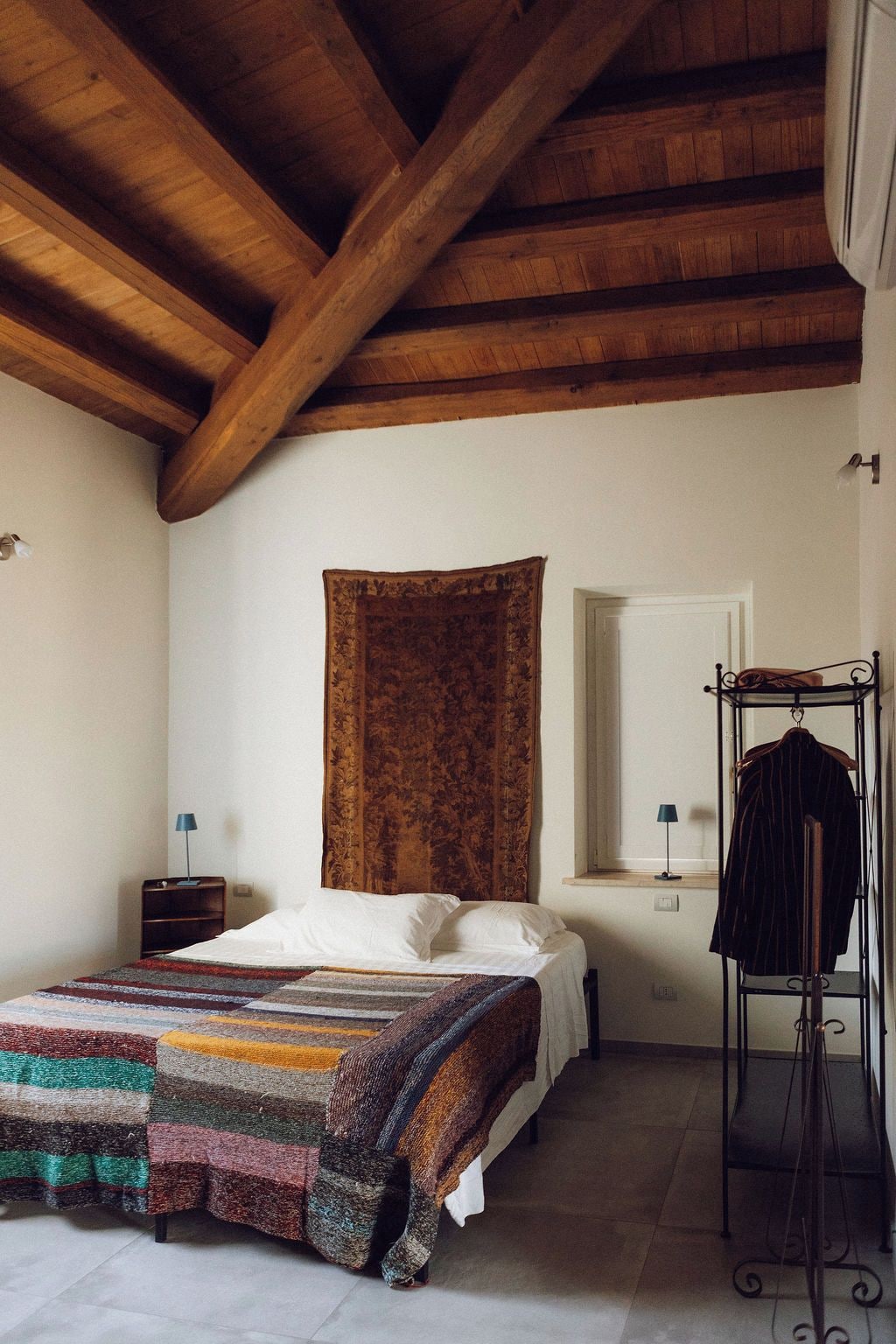 Un dormitorio con techo de madera, una cama con manta a rayas, un armario colgado y una ventana con cortinas.
