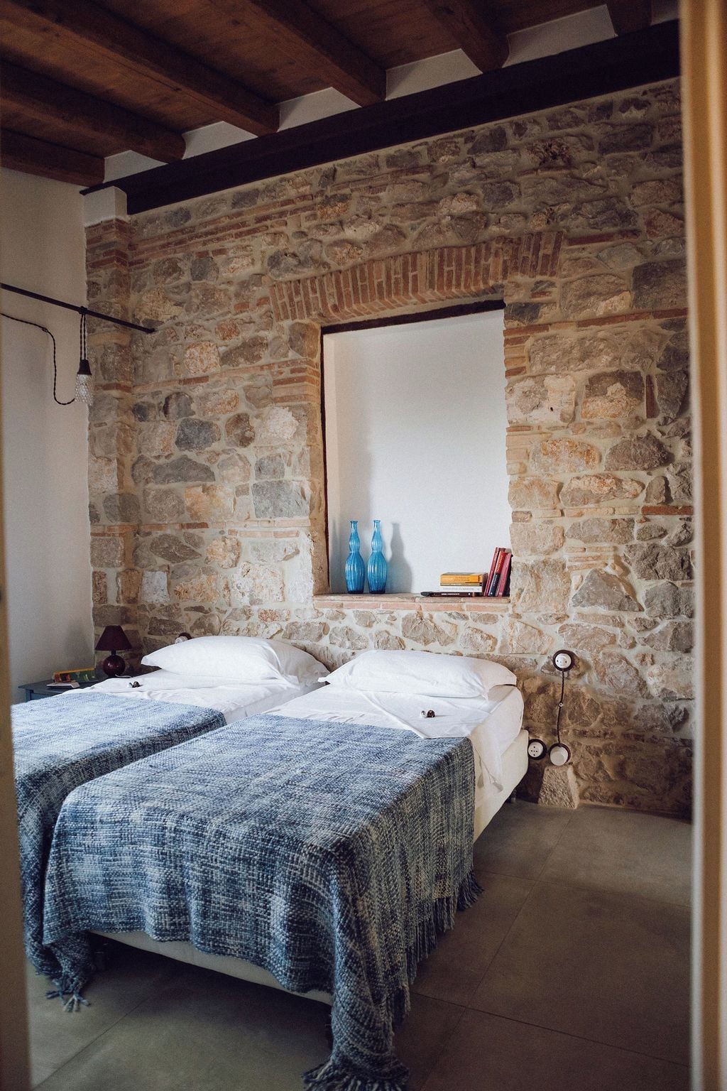 A bedroom with two beds, stone walls, a wooden ceiling, a mirror, and a shelf with books and bottles.