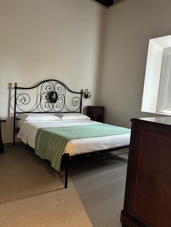 A single bed with a metal frame and a green-striped bedspread, a wooden nightstand, a wall-mounted lamp, and a wooden dresser.
