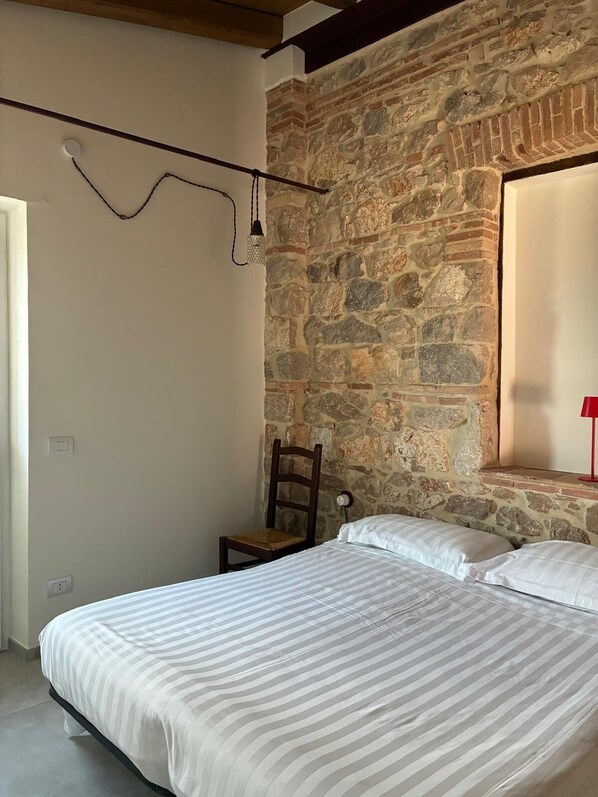A bedroom with a stone wall, a bed with striped bedding, a wooden chair, and a red lamp.