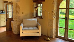 Ein Zimmer mit Holzbalkendecke, einer Couch, einer Stehlampe, einem Fenster mit Blick auf Grünflächen und einer Tür zu einem anderen Raum.