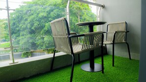 Un balcón con dos sillones de mimbre y una mesa redonda pequeña, sobre un piso de césped artificial verde, con vistas a un paisaje verde exuberante.