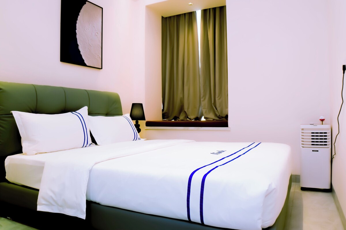 A hotel room with a bed, a lamp, a window with curtains, and a wall-mounted air conditioner.