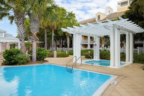 Une piscine agrémentée d’une pergola blanche, de palmiers et d’un espace clos avec un bâtiment visible au fond.