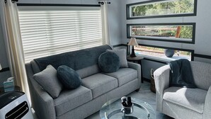 En moderne stue med en grå sofa, et rundt glasbord og udsigt til en skov gennem vinduerne.
