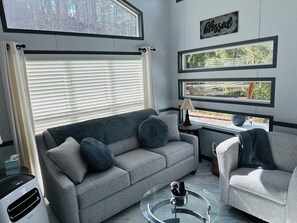 Ein modernes Wohnzimmer mit einem grauen Sofa, einem runden Glastisch und Blick auf einen Wald durch die Fenster.