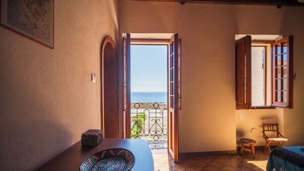 Un umbral abierto que da a un balcón con vista al mar, una mesa de madera con una cesta tejida y una silla.