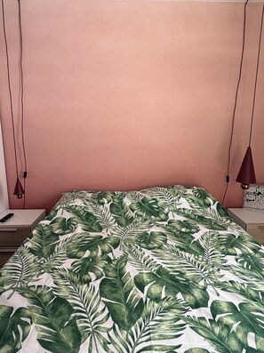 A bed with a tropical leaf pattern bedspread, a bedside table with a lamp, and a wall-mounted power outlet.