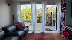 A living room with a black leather sofa, a red rug, a bookshelf, and a sliding glass door leading to a patio with plants and a view of buildings.