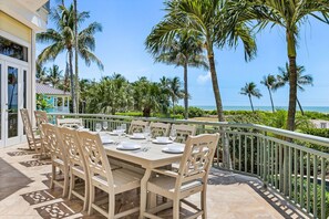 A dining area with a table set for six, surrounded by chairs, overlooking a beach with palm trees and a clear sky. - 5 Bedroom / 5 Bath Luxury Estate (Captiva)