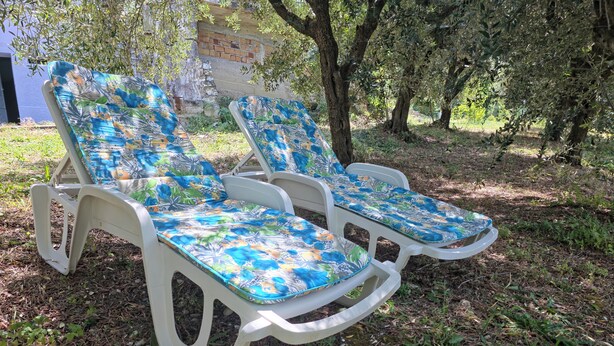 Dos sillones blancos de plástico con cojines estampados, situados en un césped con árboles y un muro de ladrillo al fondo.