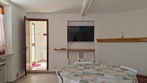 Una habitación con una mesa de comedor, sillas blancas, un televisor montado en la pared y una puerta que da a otra estancia.