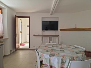 A room with a dining table, white chairs, a TV mounted on the wall, and a door leading to another room. - Casa Vacanze Merion (Arcevia)